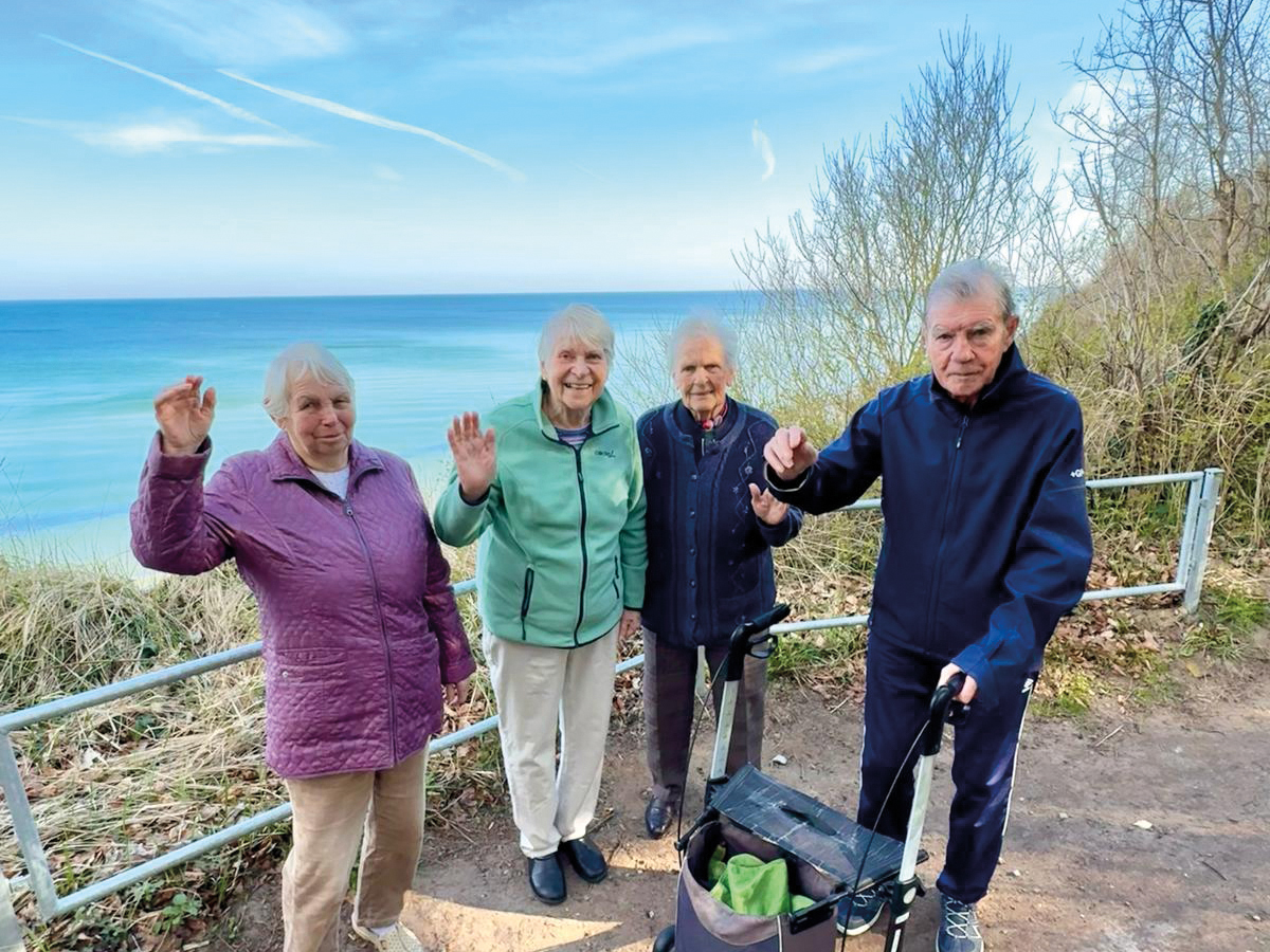 Vier Bewohner stehen auf der Aussichtsebene vor der Steilküste und winken in die Kamera. Im Hinergrund ist die blaue Ostsee zu sehen.
