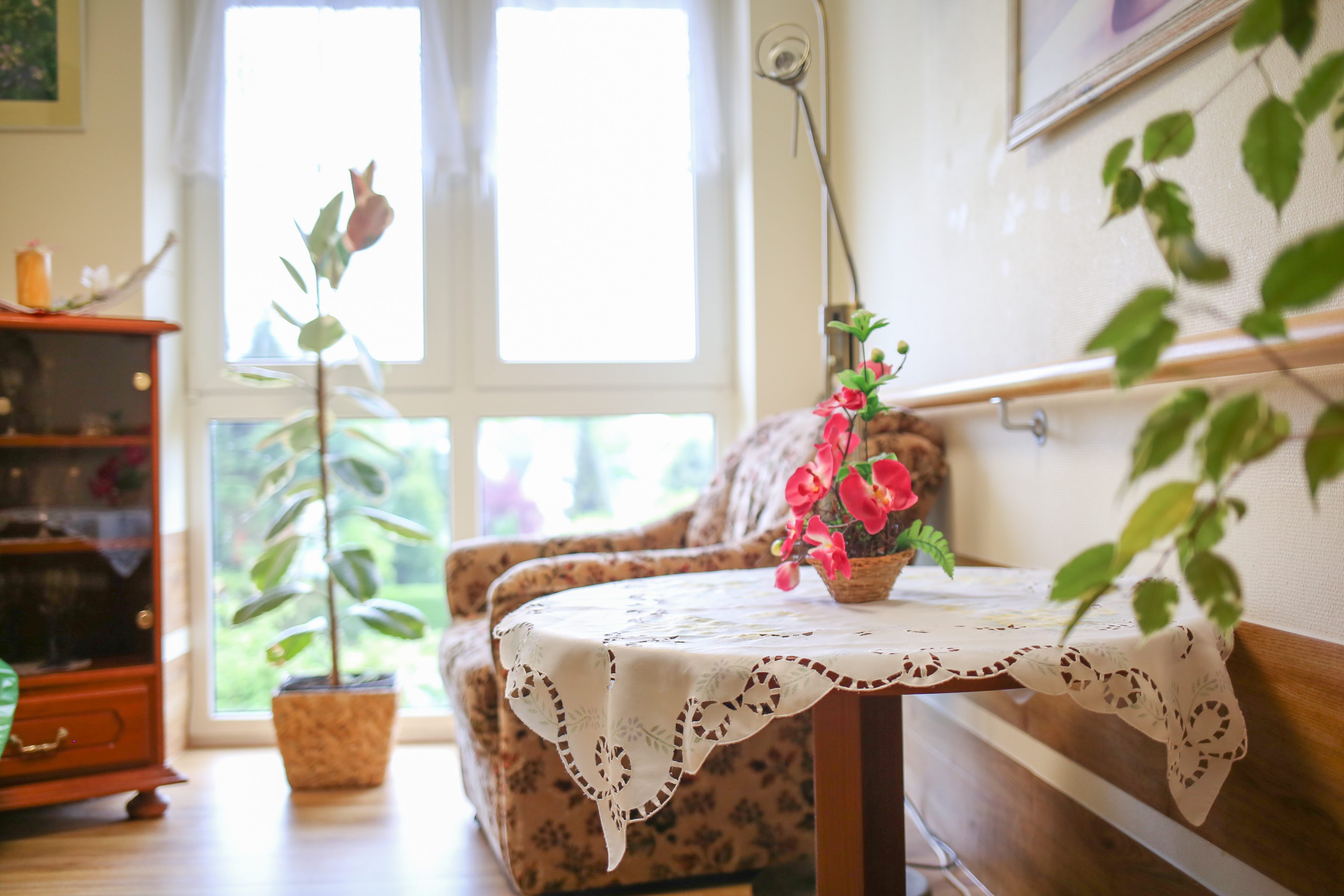 Ein Tisch mit Blume darauf in einem hellen Raum, im Hintergrund ein großes Fenster mit Grünpflanze davor