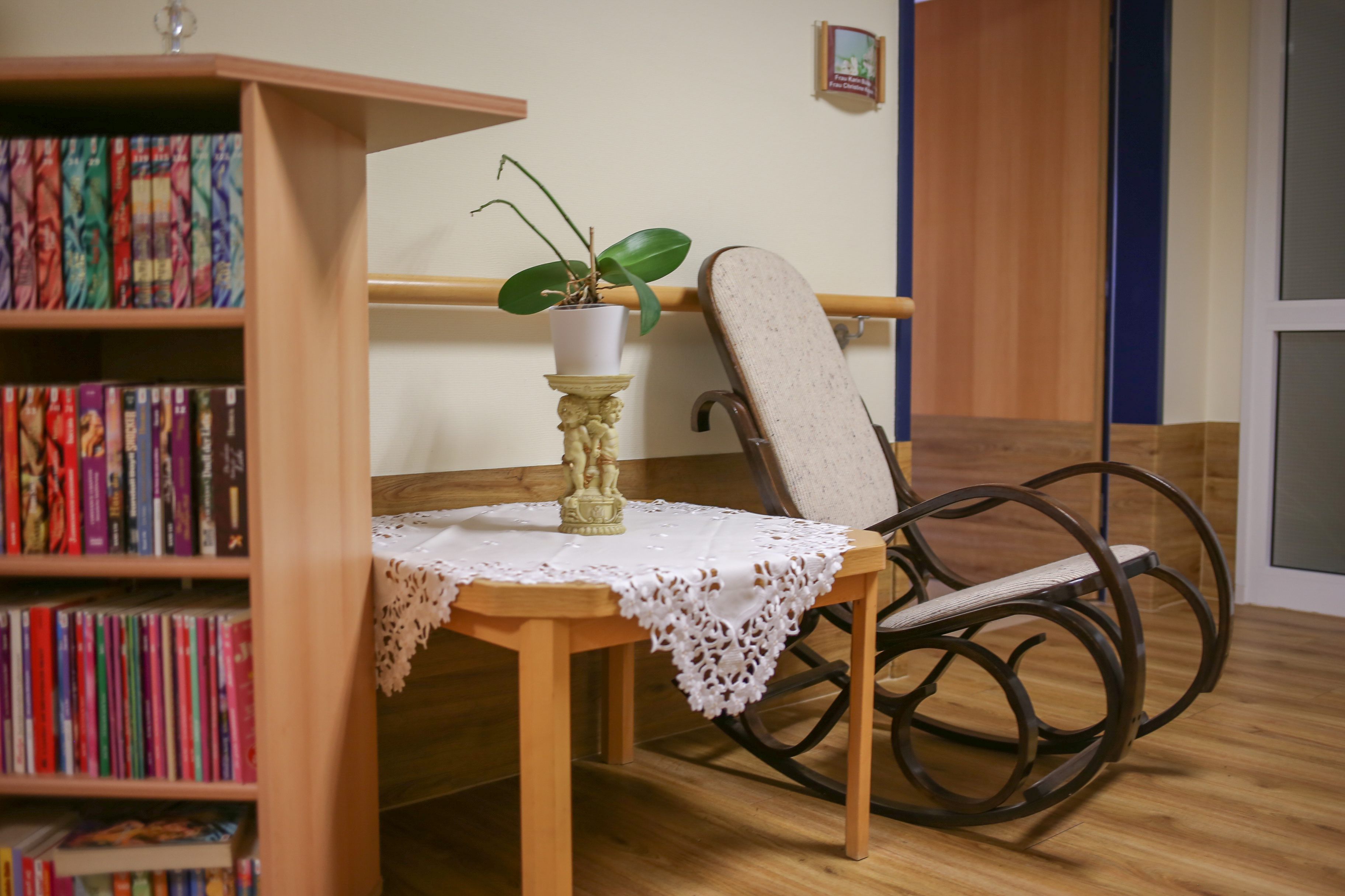 Ein Bücherregal als kleiner Raumtrenner, dahinter ein kleiner Tisch mit Grünpflanze and am Tisch ein Schaukelstuhl