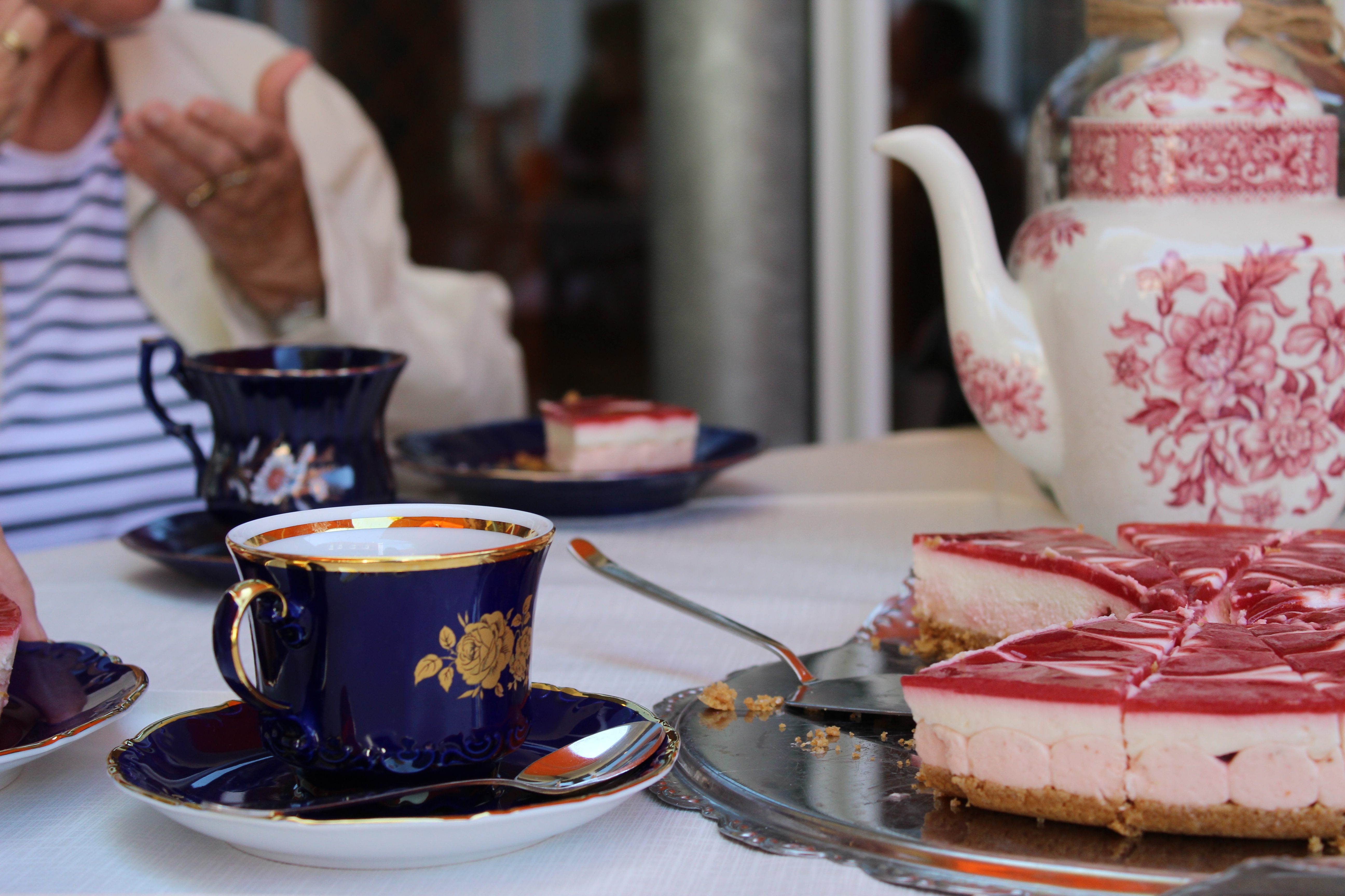 Ein gedeckter Tisch mit Kaffeekanne, Kaffeetassen und leckerer Erdbeertorte, im Hintergrund eine ältere Dame.